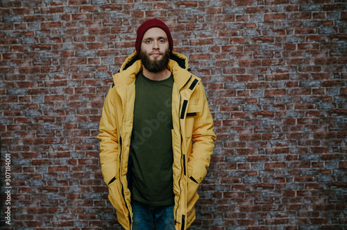City portrait of handsome hipster guy with beard wearing a blank winter yellow jacket and red hat standing on a brick wall background. Empty space for your logo or design. Mockup for print. © San4ezz007