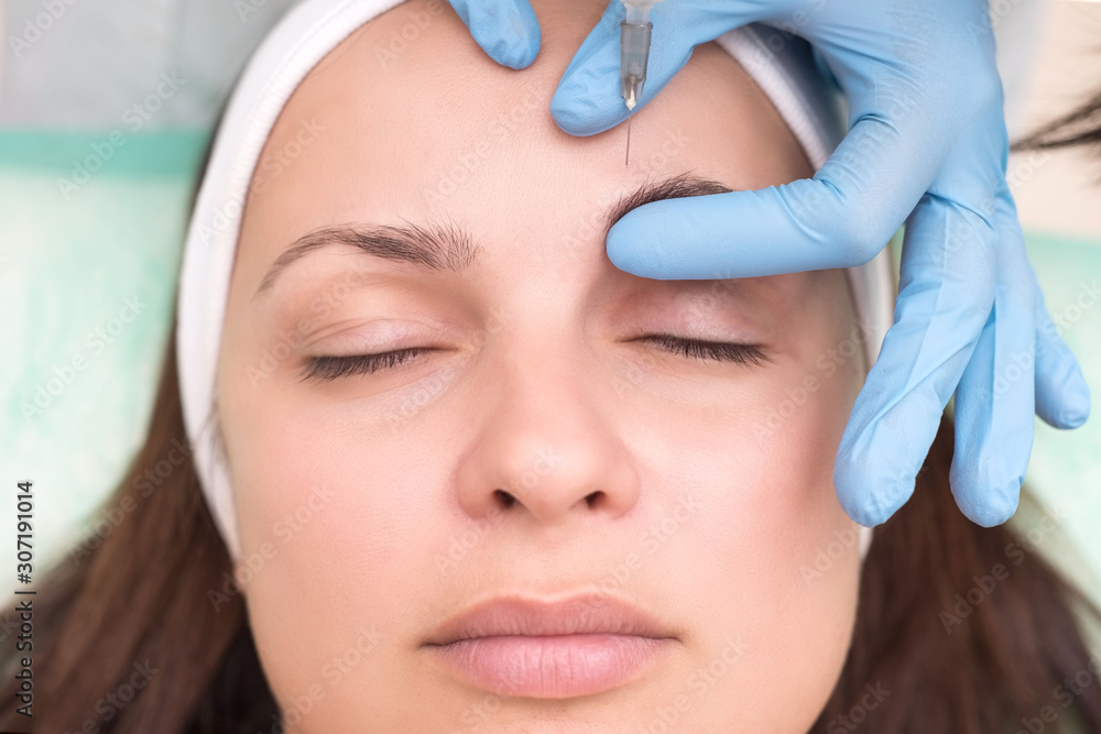 injection into the skin of the face of a young woman, close-up. Beautician with a syringe with a needle introduces a tool to remove wrinkles on the face in the eyebrow area.