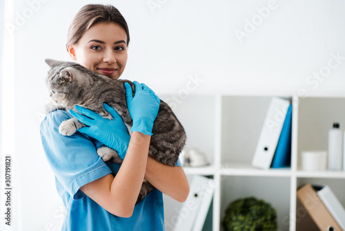 smiling veterinarian looking at camera while holding grey tabby cat on hands photo