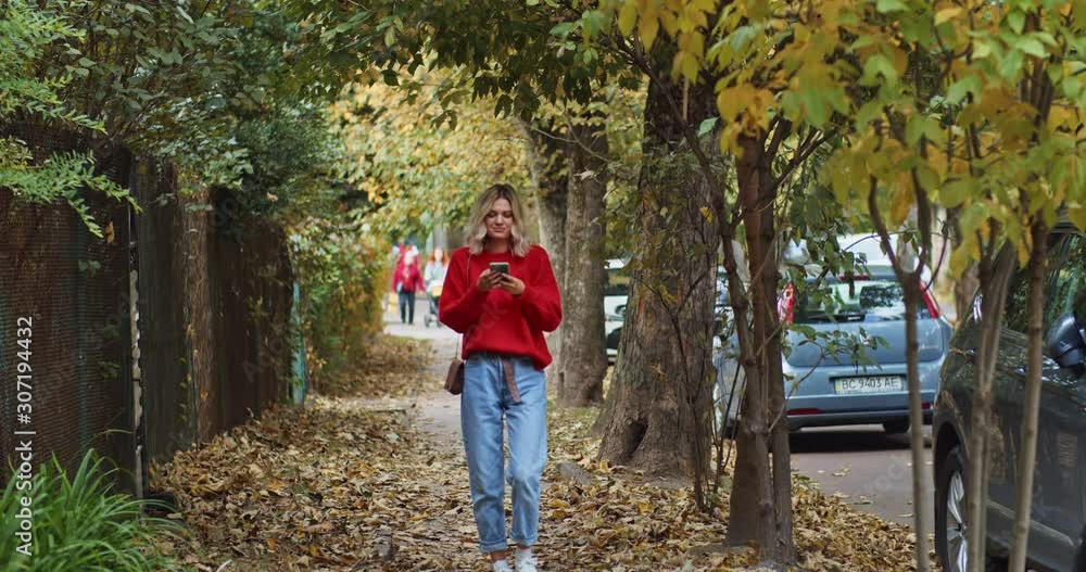 Handsome pretty caucasian blonde woman using smartphone walking outside on the street smiling looking at mobile screen. Enjoying autumn city. Online communication. Shopping app notification. Female Handsome pretty caucasian blonde woman using smartphone walking outside on the street smiling looking at mobile screen. Enjoying autumn city. Online communication. Shopping app notification. Female