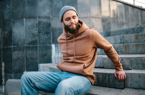 City portrait of handsome hipster guy with beard wearing brown blank hoodie or hoody and hat with space for your logo or design. Mockup for print © San4ezz007