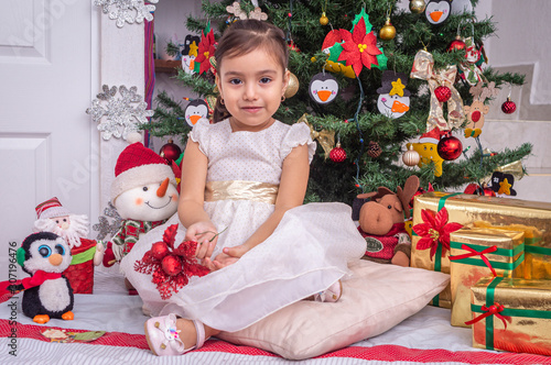 Smiling 3 year old Mexican girl sitting in front of Christmas tree