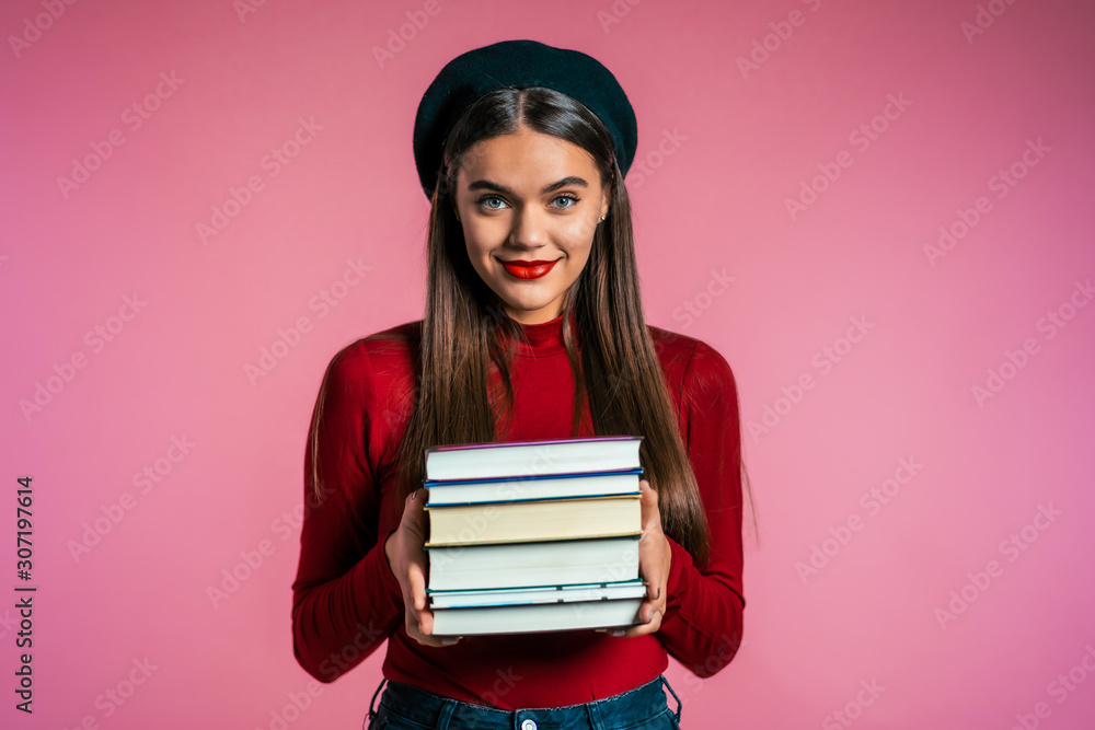 Pretty student on pink background in studio holds stack of university books from library. Girl in hat smiles, she is happy to graduate. Pretty student on pink background in studio holds stack of university books from library. Girl in hat smiles, she is happy to graduate.