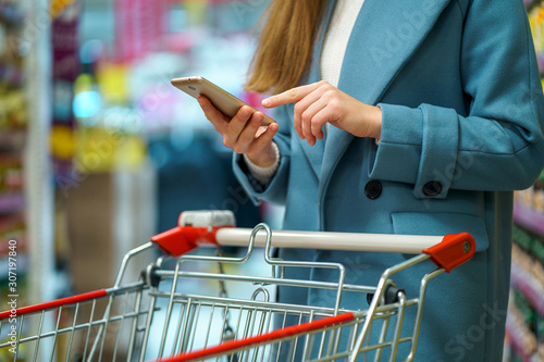 Woman buyer with cart in the shop aisle with grocery list on smartphone during shopping food photo