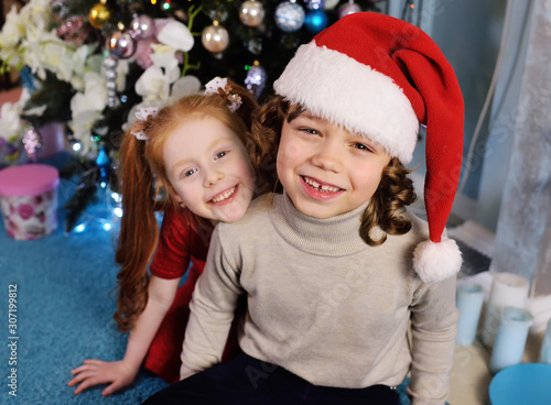 small children - a boy with curly hair in a Santa hat and a girl with red hair smiling and hugging on the background of a Christmas tree.