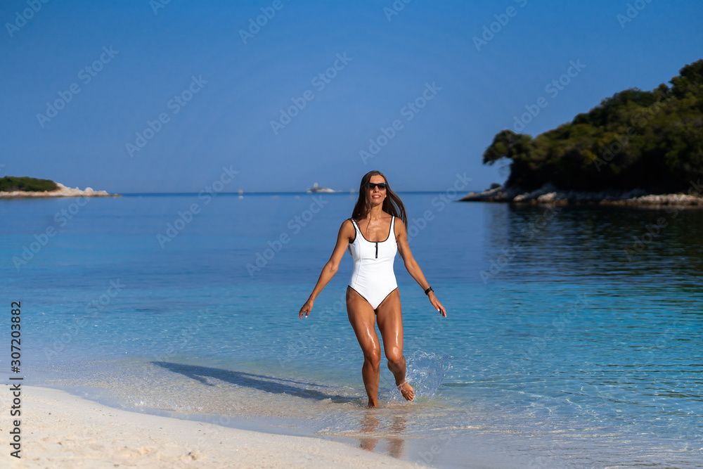 Fototapeta premium Young girl running on beach. Athletic woman jogging in trendy sexy white bodysuit enjoying the sun exercising. Healthy lifestyle. Fun walk along the shore. Perfect fitness body shapes and tan skin