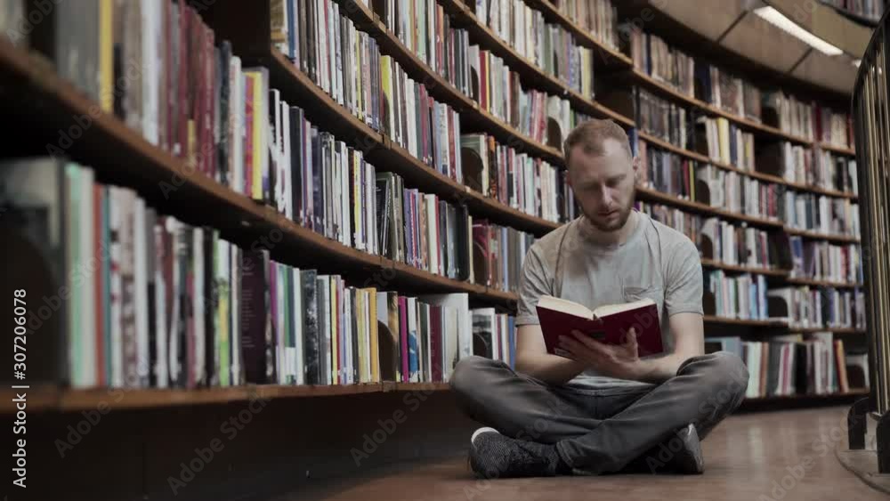 A male student is sitting on the floor among the bookshelves of the university library and enthusiastically studying, reading a book or textbook. A male student is sitting on the floor among the bookshelves of the university library and enthusiastically studying, reading a book or textbook.