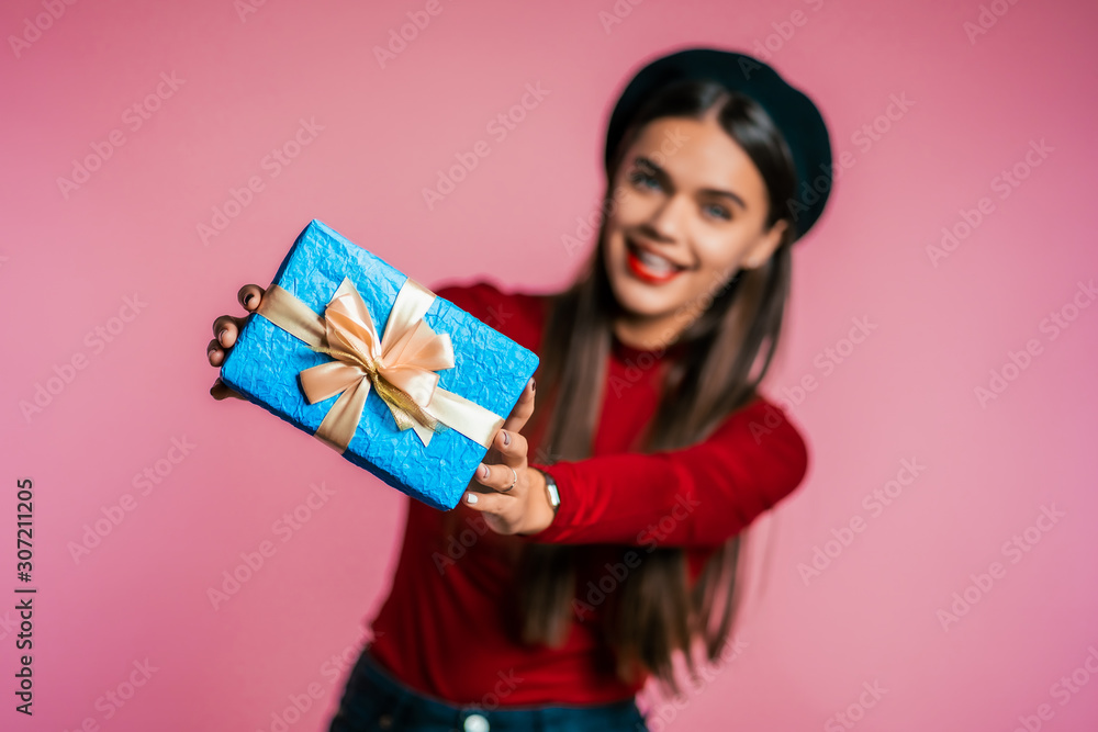 Pretty woman gives gift and hands it to the camera. She is happy, smiling. Girl on pink background. Positive holiday shot. Pretty woman gives gift and hands it to the camera. She is happy, smiling. Girl on pink background. Positive holiday shot.