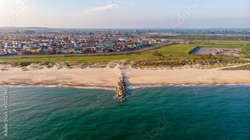 An aerial view of a sandy beach with crystal green water and rocky groyne (breakwater) along a coastal urban area in the background under a blue sky and white clouds
