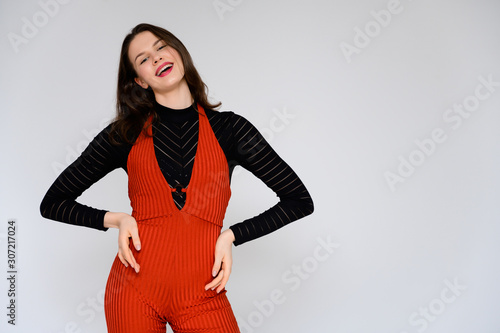 Concept adult girl on a white background. A photo of a pretty brunette girl in red trousers and a black sweater smiles and shows different emotions in different poses right in front of the camera. © Вячеслав Чичаев