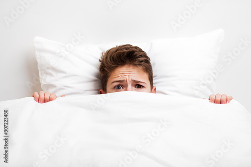 Scared peeking child boy lying bed covering face with blanket