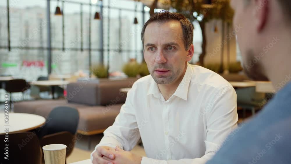 Tilt up of middle aged businessman sitting in cafe and discussing cooperation with business partner over coffee, open laptop and notepad on table Tilt up of middle aged businessman sitting in cafe and discussing cooperation with business partner over coffee, open laptop and notepad on table