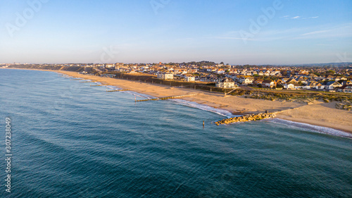 An aerial view of a sandy beach with crystal green water and rocky groyne (breakwater) along a coastal urban area in the background under a blue sky and white clouds