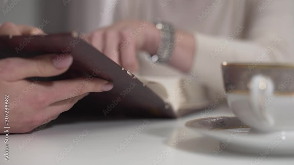 Close-up of female Caucasian hand holding photo album. Older unrecognizable woman turning pages a the background. Mother and daughter sharing memories indoors. Close-up of female Caucasian hand holding photo album. Older unrecognizable woman turning pages a the background. Mother and daughter sharing memories indoors.