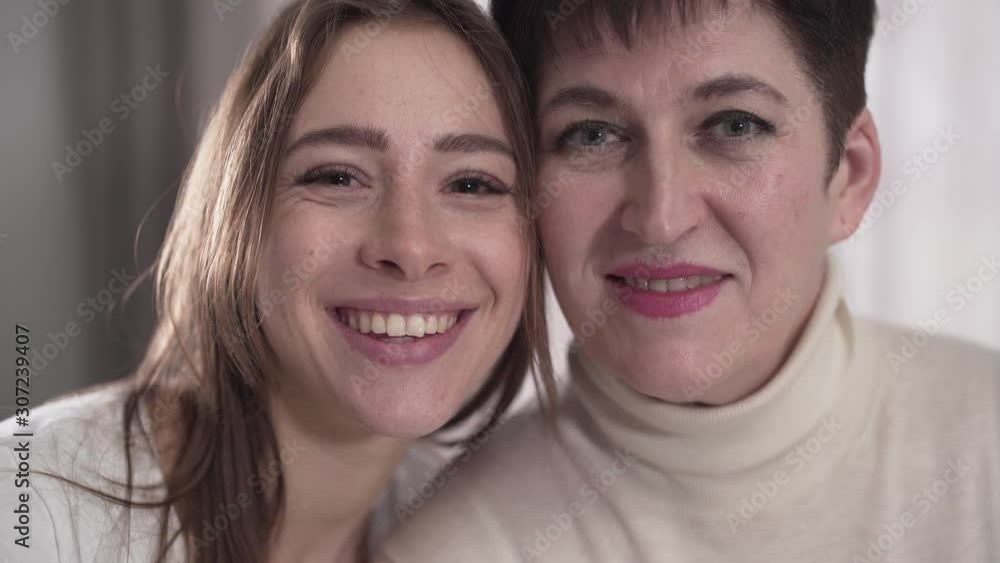 Portrait of happy Caucasian mother and daughter looking at camera and smiling. Cheerful young woman and her young-looking senior mom posing indoors. Happiness, bonding, good relationship. Portrait of happy Caucasian mother and daughter looking at camera and smiling. Cheerful young woman and her young-looking senior mom posing indoors. Happiness, bonding, good relationship.
