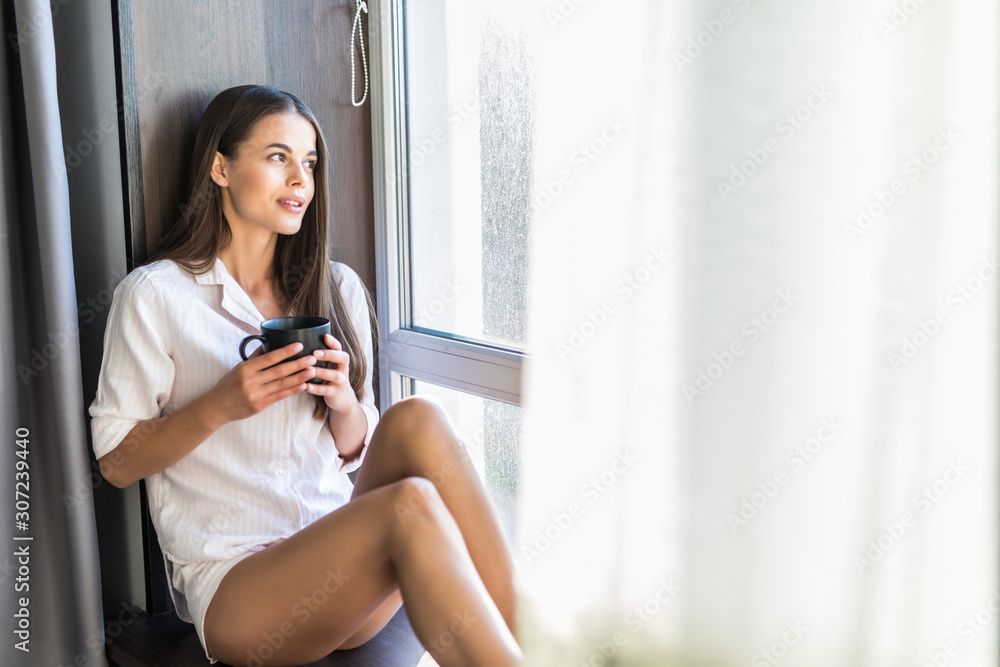 Beautiful young woman drinking coffee and looking through a window Beautiful young woman drinking coffee and looking through a window