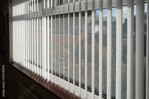 White vertical blinds slats hanging in front of double glazed white frame window. The slats have no cords at the bottom. The focus is shallow.