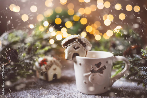 Miniature gingerbread house on a chpshka against the background of a Christmas tree of fire garland. Snowfall effect.