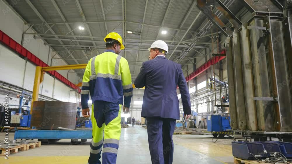 Follow footage of caucasian businessman or engineer and black worker in hard hat walking through factory facility talking. Inspector visiting plant