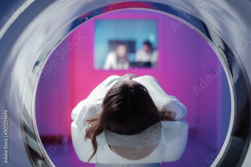 The girl patient is lying in the tomograph and waiting for a scan. Three doctors from the exam room look at the pictures photo