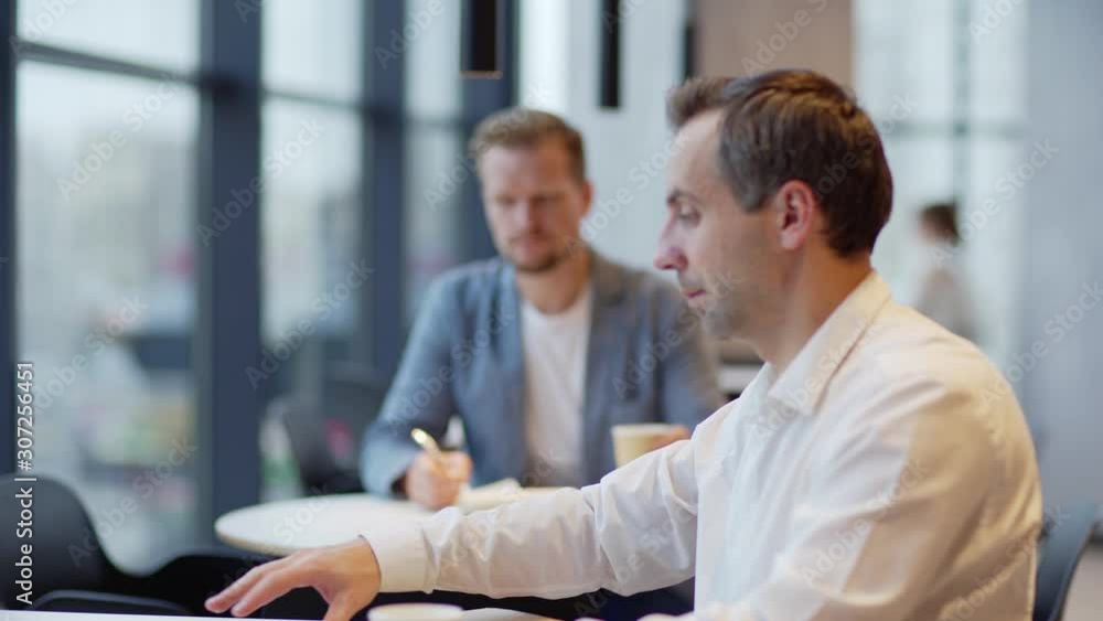 Camera moving from middle aged man writing in notepad and drinking coffee to businessman opening laptop computer and typing sitting at table in cafe, rack focus Camera moving from middle aged man writing in notepad and drinking coffee to businessman opening laptop computer and typing sitting at table in cafe, rack focus