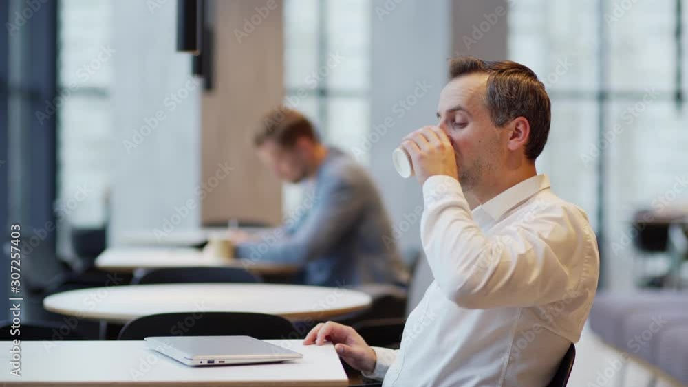 Side view medium shot of businessman stretching arms and drinking coffee to cheer up in morning, then opening laptop computer and typing with smile on his face sitting at table in cafe Side view medium shot of businessman stretching arms and drinking coffee to cheer up in morning, then opening laptop computer and typing with smile on his face sitting at table in cafe