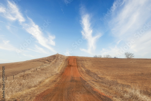 A red dirt road going uphill dividing fenced pasture land in a brown springgtime landscape in South Dakota USA