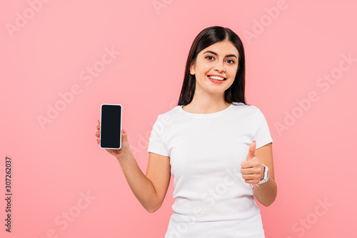 smiling pretty brunette girl holding smartphone with blank screen and showing thumb up isolated on pink