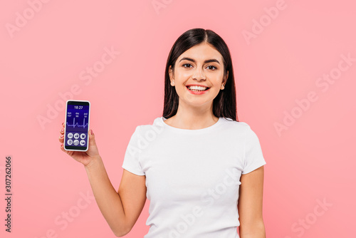  smiling pretty brunette girl holding smartphone with healthcare app isolated on pink