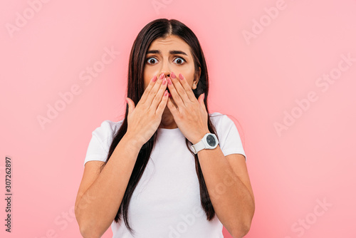 scared pretty brunette girl covering mouth with hands isolated on pink photo