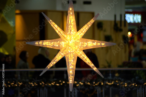 Golden glittering eight-pointed star of Bethlehem in a building with New Year decorations and people silhouettes. Background for Christmas.