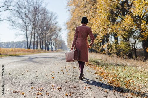 Young redhead lady woman in pink vintage coat and hat with suitcase in retro style walking away along a park road with golden yellow autumnal trees. Outdoor autumn garden relaxation, travel concept