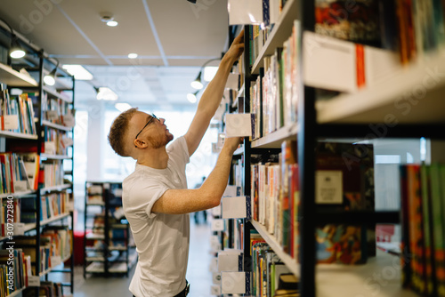 Focused guy reaching for book on top shelf