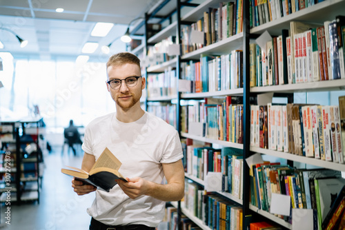 Adult guy holding opened book and looking at camera in library
