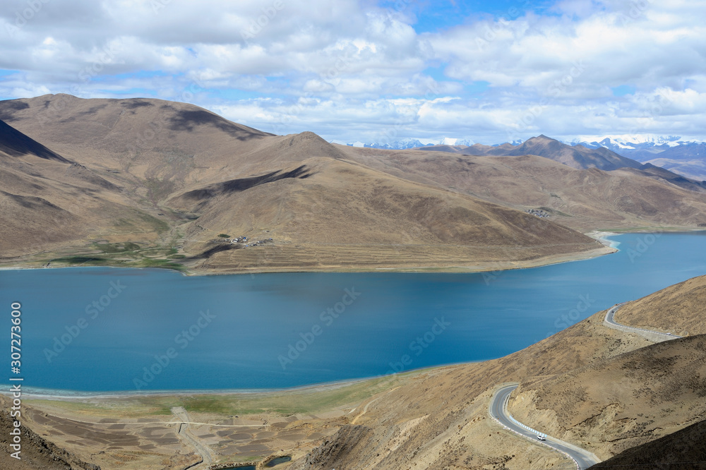 Nestled among the flanks of the dull gray and green mountains, sacred Lake Yamdrok glows turquoise when the sun is shining in Tibetan Plateau in Brahmaputra Valley of the Tibet Autonomous Region of Ch