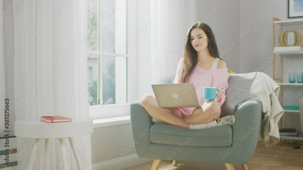 Portrait of Young Brunette Works on Laptop Computer while Holding Cup with Hot Beverage, and Drinking From it With Pleasure. Gorgeous Girl Wearing Pink Sweater, Spends Relaxing Time in Living Room Portrait of Young Brunette Works on Laptop Computer while Holding Cup with Hot Beverage, and Drinking From it With Pleasure. Gorgeous Girl Wearing Pink Sweater, Spends Relaxing Time in Living Room