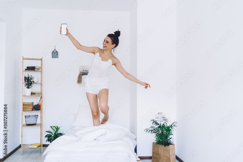 Carefree woman with smartphone in pyjamas dancing on bed Carefree woman with smartphone in pyjamas dancing on bed