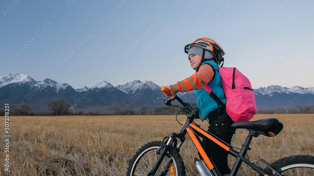 One caucasian children walk with bike in wheat field. Little girl walking black orange cycle on background of beautiful snowy mountains. Biker stand with backpack and helmet. Mountain bike hardtail.