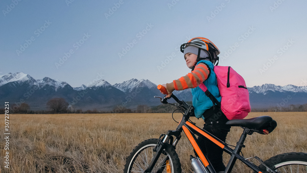 One caucasian children walk with bike in wheat field. Little girl walking black orange cycle on background of beautiful snowy mountains. Biker stand with backpack and helmet. Mountain bike hardtail. One caucasian children walk with bike in wheat field. Little girl walking black orange cycle on background of beautiful snowy mountains. Biker stand with backpack and helmet. Mountain bike hardtail.
