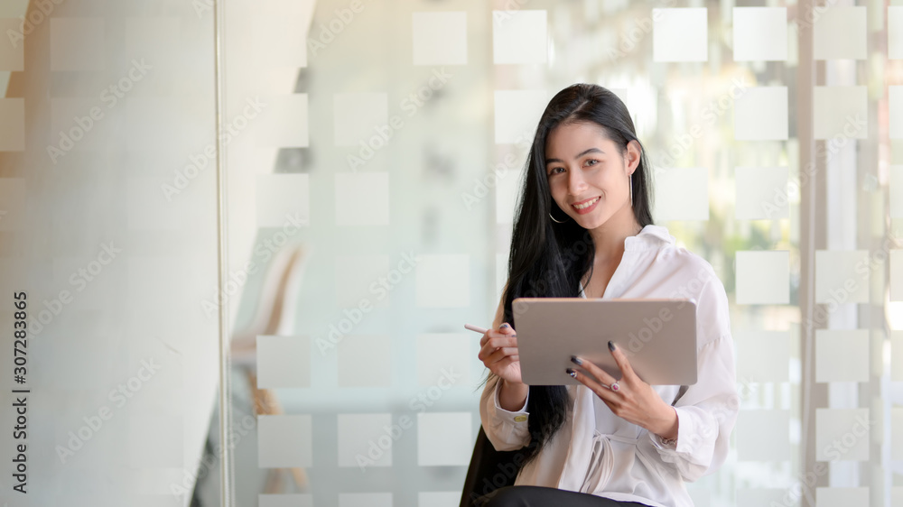 Portrait of young beautiful graphic designer working on her project with tablet and looking at the camera while standing in modern office Portrait of young beautiful graphic designer working on her project with tablet and looking at the camera while standing in modern office