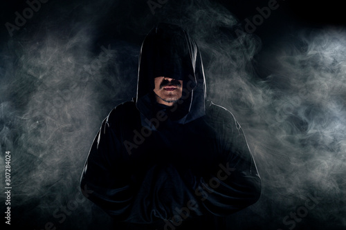 Man dressed in a dark robe looking like a cult leader on a smoky or foggy background.  He looks like a creepy evil villain