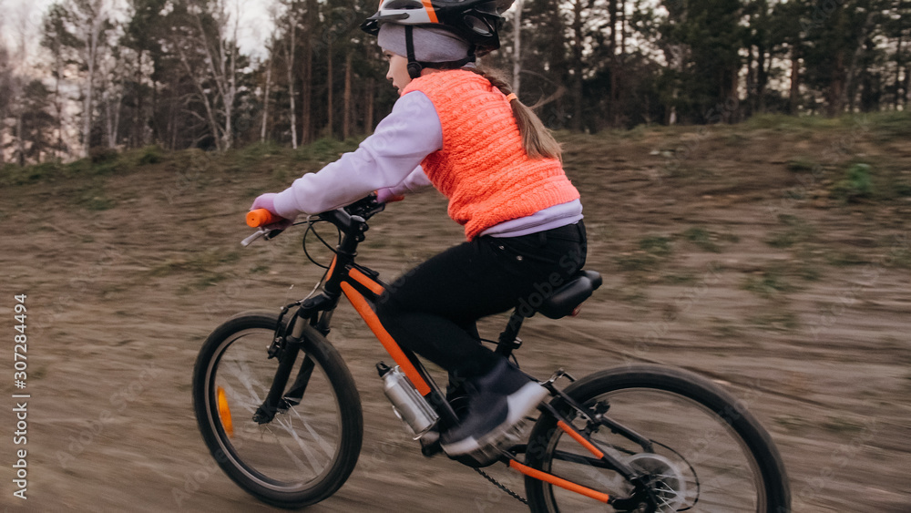 Naklejka premium One caucasian children rides bike road track in dirt park. Girl riding black orange cycle in racetrack. Kid goes do bicycle sports. Biker motion ride with backpack and helmet. Mountain bike hardtail.