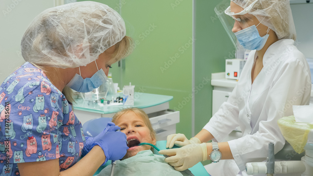 Child with a mother at a dentist's reception. The girl lies in the chair, behind her mother. The doctor works with an assistant. Procedure for drilling a tooth. Setting a temporary seal. Using the x Child with a mother at a dentist's reception. The girl lies in the chair, behind her mother. The doctor works with an assistant. Procedure for drilling a tooth. Setting a temporary seal. Using the x