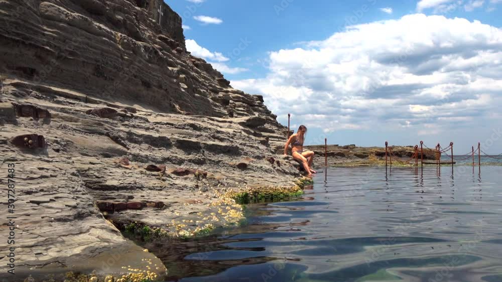 A girl in a bikini lowers her foot in the cold lake under the rock A girl in a bikini lowers her foot in the cold lake under the rock