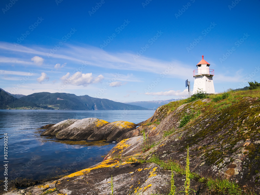 White lighthouse tower with red top and moss covered rocky shore of the Sognefjord in Norway on a sunny day, with mountains, blue sky and fluffy white clouds. White lighthouse tower with red top and moss covered rocky shore of the Sognefjord in Norway on a sunny day, with mountains, blue sky and fluffy white clouds.