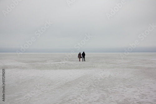 A couple a man and a woman silhouette walk together in the distance on the ice of a frozen snow covered endless winter lake to horizon