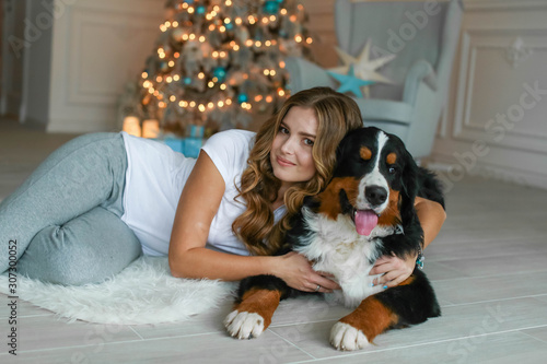 A beautiful woman with long hair lies on a rug against the background of a Christmas tree and next to her lies her big dog.