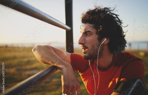 Side view of an exhausted young male athlete listening to music on earphones leaning on bars in gym park looking away into the sunset photo