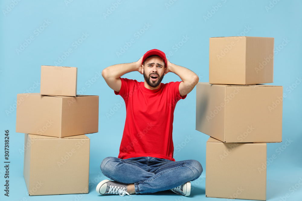 Delivery sad man in red uniform isolated on blue background, studio portrait. Male employee in cap t-shirt print working as courier dealer hold empty cardboard box. Service concept. Mock up copy space Delivery sad man in red uniform isolated on blue background, studio portrait. Male employee in cap t-shirt print working as courier dealer hold empty cardboard box. Service concept. Mock up copy space