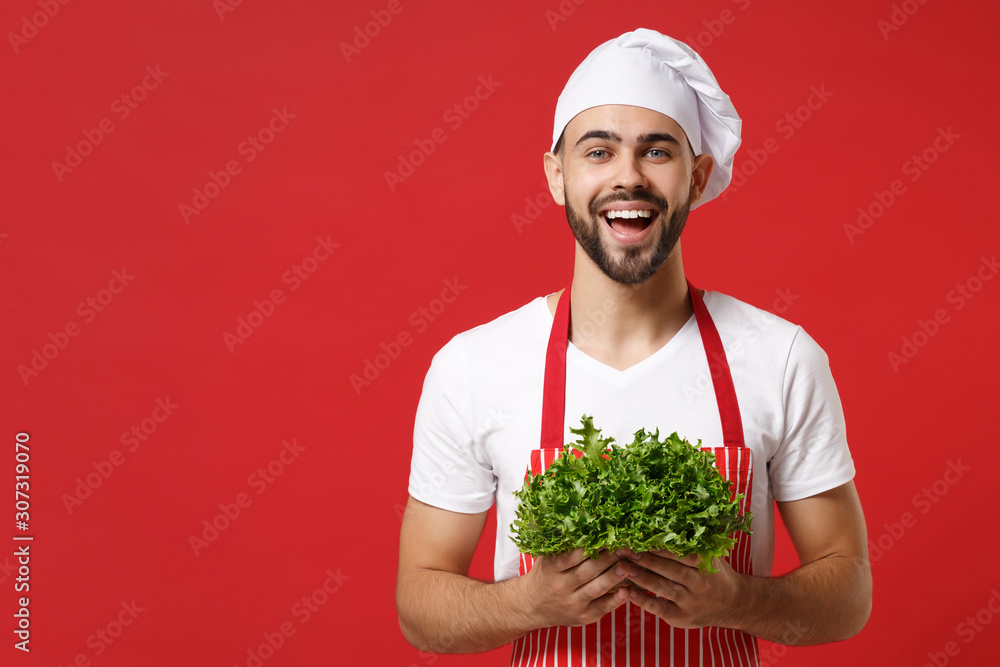 Laughing young bearded male chef cook or baker man in striped apron white t-shirt toque chefs hat isolated on red background. Cooking food concept. Mock up copy space. Hold green fresh salad leaves. Laughing young bearded male chef cook or baker man in striped apron white t-shirt toque chefs hat isolated on red background. Cooking food concept. Mock up copy space. Hold green fresh salad leaves.